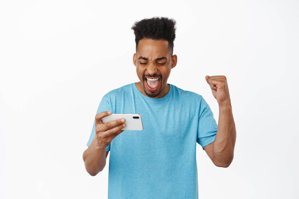 Portrait of happy african american man rejoicing from winning social media contests on mobile phone, looking at smartphone horizontal and fist pump, watching stream on phone, white background